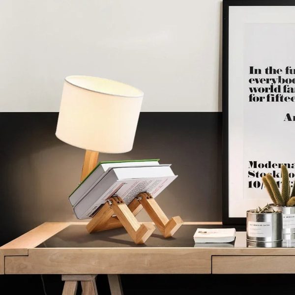 Luminária de mesa com base de madeira em forma de robô, com abajur branco, colocada numa secretária de escritório moderna com quadro e plantas ao fundo.