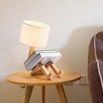 Luminária de mesa em forma de figura humana com cabeça de abajur, apoiada em uma mesa de madeira clara ao lado de um sofá castanho.
