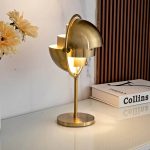 Candeeiro de mesa com design moderno em metal dourado, com duas lâmpadas semicirculares e base circular, colocado numa mesa branca ao lado de livros e flores.