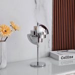 Candeeiro de mesa em metal com design futurista, base circular e acabamento cromado, colocado numa secretária branca com livros e flores amarelas ao lado.
