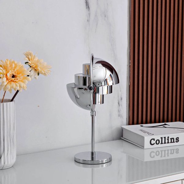 Candeeiro de mesa em metal com design futurista, base circular e acabamento cromado, colocado numa secretária branca com livros e flores amarelas ao lado.
