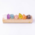Vários cristais de diferentes cores e tamanhos dispostos numa base de madeira, incluindo cristal transparente, roxo, azul, rosa, amarelo, laranja e roxo escuro.