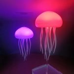 Luminária de medusa com cabeça iluminada em rosa e corpo em tentáculos ondulados, emitindo luz suave em um ambiente escuro.