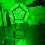 Candeeiro de mesa com estrutura hexagonal e luzes verdes LED, em ambiente de sala com livros e objetos decorativos ao fundo.