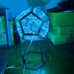 Luminária de mesa com estrutura geométrica de várias faces, iluminada por LEDs azuis, em ambiente de escritório com livros e caixas verdes ao fundo.
