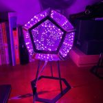 Luminária de mesa com estrutura geométrica em forma de dodecaedro, iluminada com luzes LED roxas, em ambiente escuro, com livros e objetos ao fundo.