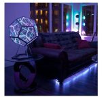 Luminária de design geométrico com luzes LED azuis e roxas, colocada ao lado de um sofá de veludo preto em sala com iluminação ambiente colorida.