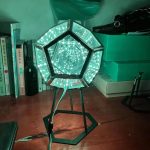 Lâmpada de mesa com estrutura hexagonal e luzes LED verdes formando um efeito espiral, colocada numa mesa de estudo com livros e caixas ao fundo.