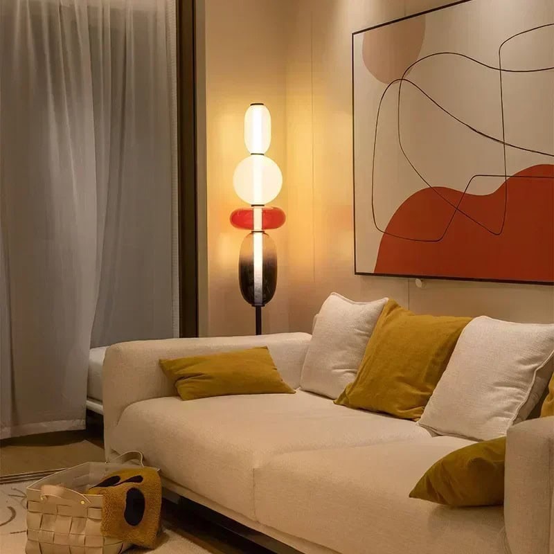 Candeeiro de mesa moderno para sala Candeeiro de mesa com design contemporâneo, base preta, duas lâmpadas brancas e uma lâmpada laranja, colocado ao lado de um sofá branco com almofadas amarelas e uma obra de arte abstrata na parede.