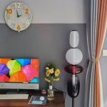Candeeiro de mesa com várias lâmpadas de diferentes tamanhos e cores, colocado ao lado de uma televisão e uma jarra com flores na sala.
