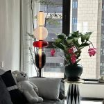 Luminária de mesa com base de vidro e estrutura de metal, decorada com elementos de vidro colorido em forma de esferas, ao lado de uma planta com flores rosas em um vaso preto.