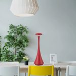 Luminária de mesa vermelha com design elegante, colocada numa mesa de escritório com plantas e dispositivo digital ao fundo.