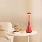 Candeeiro de mesa vermelho com design futurista, base estreita e luz suave, colocado numa mesa ao lado de uma cama com decoração minimalista.