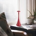 Candeeiro de mesa vermelho com design elegante, colocado numa mesa junto a uma janela com cortinas brancas, acompanhado por plantas e revistas.