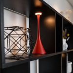 Candeeiro de mesa vermelho com design elegante e lâmpada LED, colocado numa prateleira de móvel escuro, com decoração moderna ao fundo.