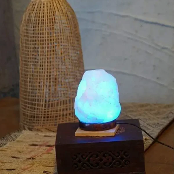 Lâmpada de sal em forma irregular com luz azul, colocada sobre base de madeira, ao lado de uma luminária de vime.