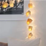 Luzes decorativas com cristais de sal em diferentes tamanhos e formas, iluminadas, penduradas ao lado de uma parede branca com quadro preto e branco.