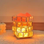 Luminária com estrutura de cubos de vidro laranja, iluminada por dentro, em ambiente acolhedor, com fundo neutro e superfície de madeira.