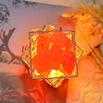 Lâmpada decorativa com cristais de quartzo laranja, iluminada, com fundo de flores e árvore ao pôr do sol.
