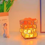 Lâmpada decorativa com cubos de vidro iluminados em tons quentes, colocada numa mesa com objetos de papel e uma figura de personagem de desenho animado ao fundo.