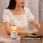 Lâmpada de sal de cristal com luz acesa, vista de lado, em ambiente de estudo, com mulher usando portátil ao fundo.