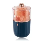 Dispositivo portátil com recipiente de sal rosa, tampa decorativa dourada e corpo azul com textura vertical, usado para condimentos ou decoração.