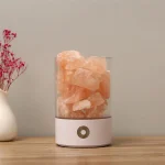 Lâmpada de sal rosa em recipiente de vidro, com luz suave, sobre mesa de madeira, ao lado de vaso com flores secas.