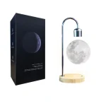 Lâmpada de lua em forma de esfera, iluminada, com suporte de metal e base de madeira, ao lado de embalagem preta com imagem da lua crescente.
