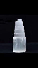 Candeeiro de selenite em forma de torre com vários níveis, iluminado, com cristal mandala na base, sobre fundo escuro.