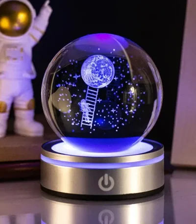 Globo de ciência com astronauta a subir uma escada até à lua, decorado com estrelas e planetas, iluminado por luz azul e roxa, em base metálica com botão de ligar.