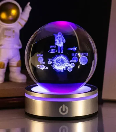 Globo de ciência com projeções de planetas, astronauta e estrelas, iluminado por luzes LED, sobre base metálica, ao lado de uma figura de astronauta de brinquedo.
