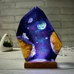 Luminária de vidro com ilustração de espaço, planetas, astronauta e estrelas, iluminada, sobre mesa de madeira com fundo de parede de tijolos.