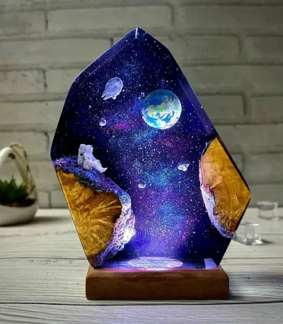 Luminária de vidro com ilustração de espaço, planetas, astronauta e estrelas, iluminada, sobre mesa de madeira com fundo de parede de tijolos.