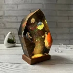 Luminária de vidro com ilustração de espaço, planetas e estrelas, iluminada, sobre mesa de madeira com fundo de parede de tijolos.