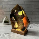 Lâmpada decorativa com design de espaço, planetas e estrelas, apoiada numa base de madeira, em ambiente com parede de tijolos cinzentos.