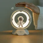 Lâmpada decorativa com design de robô e luz LED circular, sendo tocada por uma mão, com base em forma de hexágono.