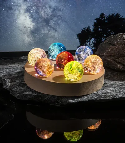 Oito bolas de vidro coloridas e iluminadas, colocadas numa base de madeira, com céu estrelado ao fundo e rochas ao redor.
