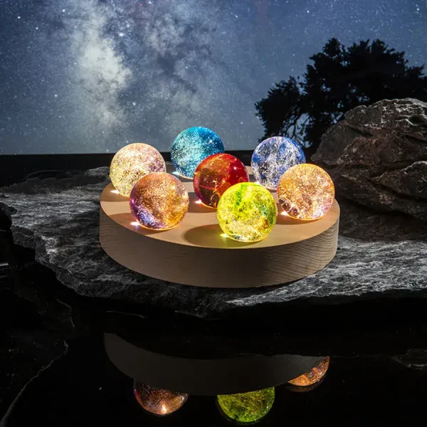 Oito bolas de vidro coloridas e iluminadas, colocadas numa base de madeira, com céu estrelado ao fundo e rochas ao redor.