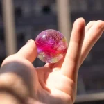 Bola de plasma transparente com cores rosa, dourado e roxo, refletindo luz solar, segurada entre dedos, com edifícios ao fundo.