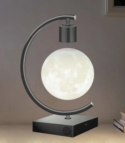 Lâmpada com base preta, estrutura metálica curva, e esfera que simula a lua, em ambiente interno com livros ao lado.