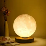 Lâmpada de lua decorativa com iluminação suave, colocada numa mesa ao lado de um vaso com flores secas e um livro aberto.