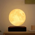Lâmpada de luz amarela com forma de lua, colocada numa base preta sobre uma mesa de madeira clara, ambiente de sala moderna.