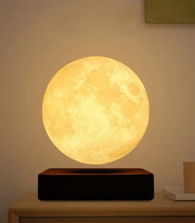 Lâmpada de luz amarela com forma de lua, colocada numa base preta sobre uma mesa de madeira clara, ambiente de sala moderna.