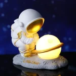 Miniatura de astronauta com capacete dourado a segurar um planeta LED brilhante, sobre base lunar com inscrição "Little Star".