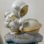 Estatueta de astronauta em pose de exploração, com capacete dourado e detalhes em ouro, apoiado numa base que simula a superfície lunar com o nome Little Star.