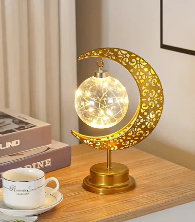 Lâmpada decorativa em forma de lua com detalhes dourados, iluminada, colocada numa mesa de madeira ao lado de livros e uma chávena.
