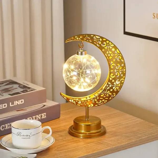 Lâmpada decorativa em forma de lua com detalhes dourados, iluminada, colocada numa mesa de madeira ao lado de livros e uma chávena.