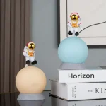 Duas figuras de astronautas em miniatura com capacetes dourados, sentados em esferas de diferentes cores, decorando uma prateleira com livros de ciência e espaço.
