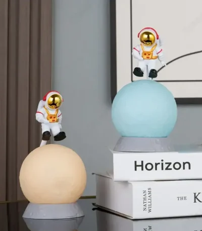 Duas figuras de astronautas em miniatura com capacetes dourados, sentados em esferas de diferentes cores, decorando uma prateleira com livros de ciência e espaço.
