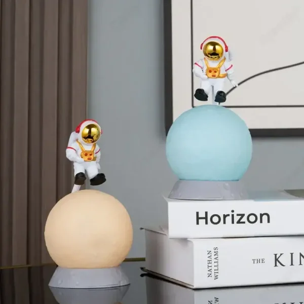 Duas figuras de astronautas em miniatura com capacetes dourados, sentados em esferas de diferentes cores, decorando uma prateleira com livros de ciência e espaço.
